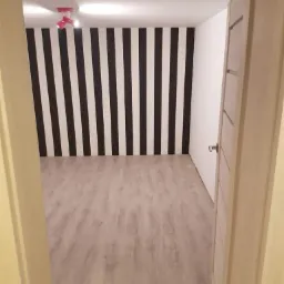 Wnętrze pokoju z podłogą imitującą drewno i ścianą w czarno-białe pionowe pasy, widok zza otwartych drzwi.