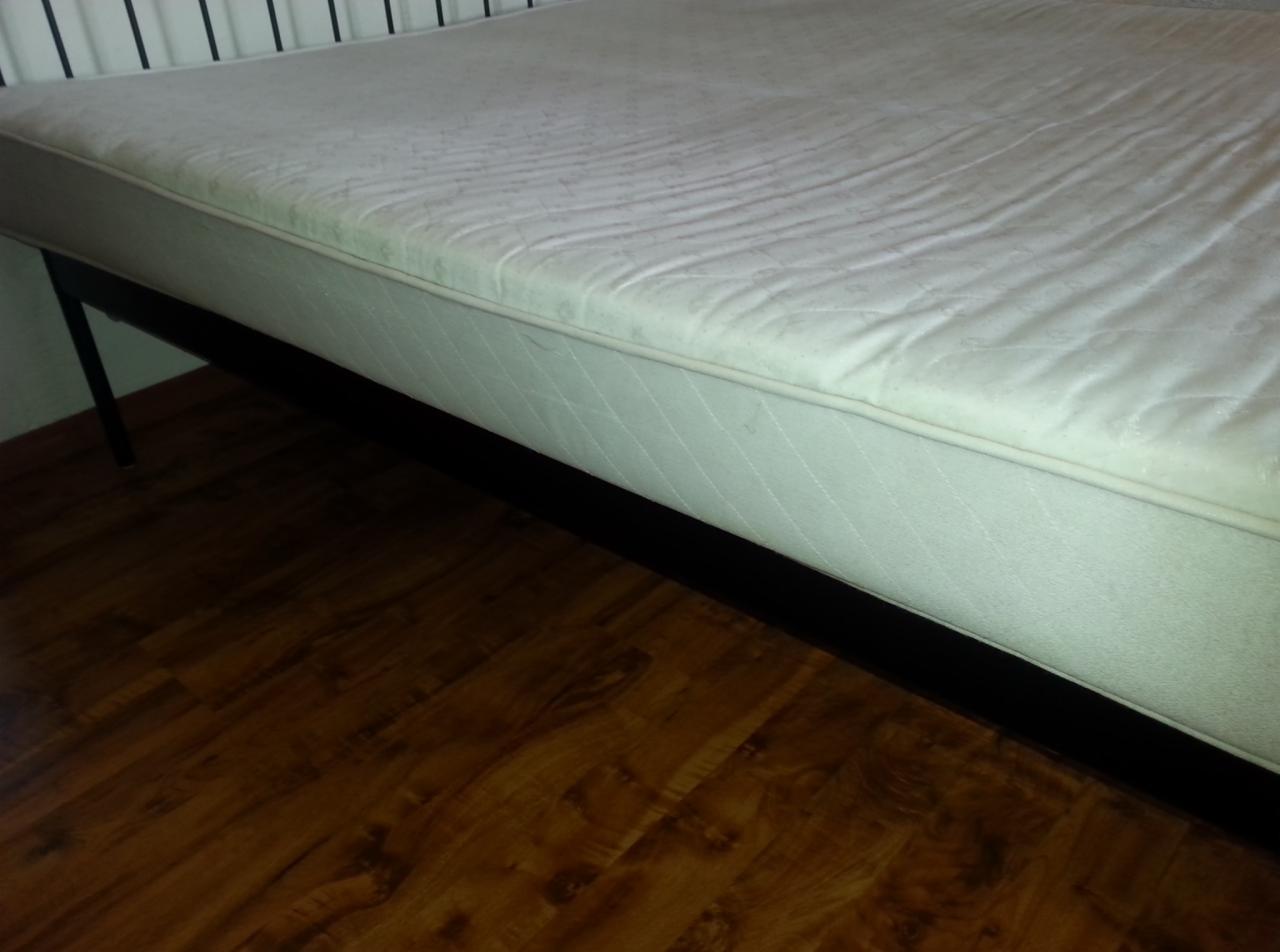 Materac z widocznymi śladami użytkowania, leżący na metalowej ramie, na tle drewnianej podłogi.