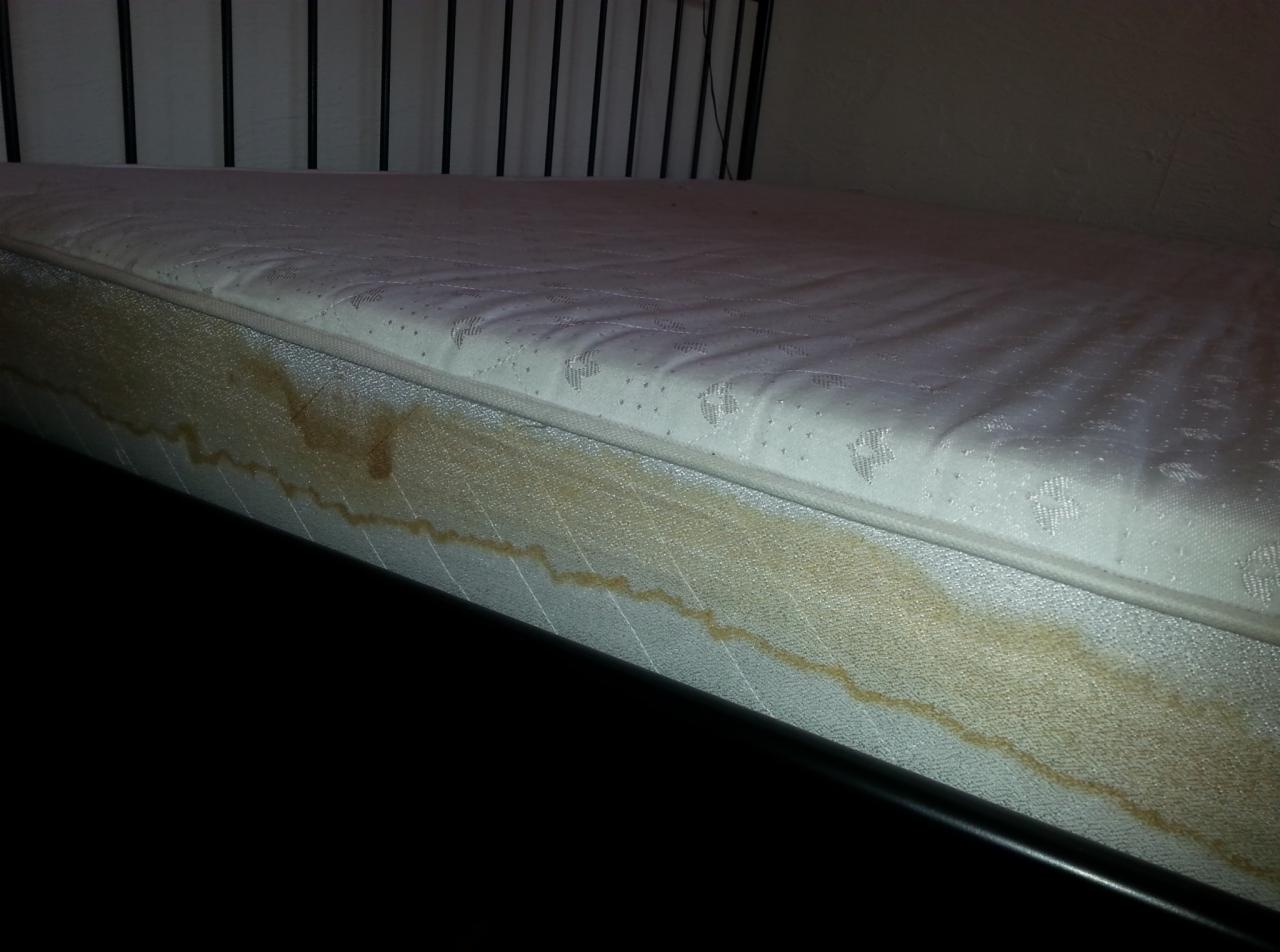 Biały materac z widocznymi, brązowymi plamami na boku, leżący na czarnej ramie łóżka, z metalowym zagłówkiem w tle.