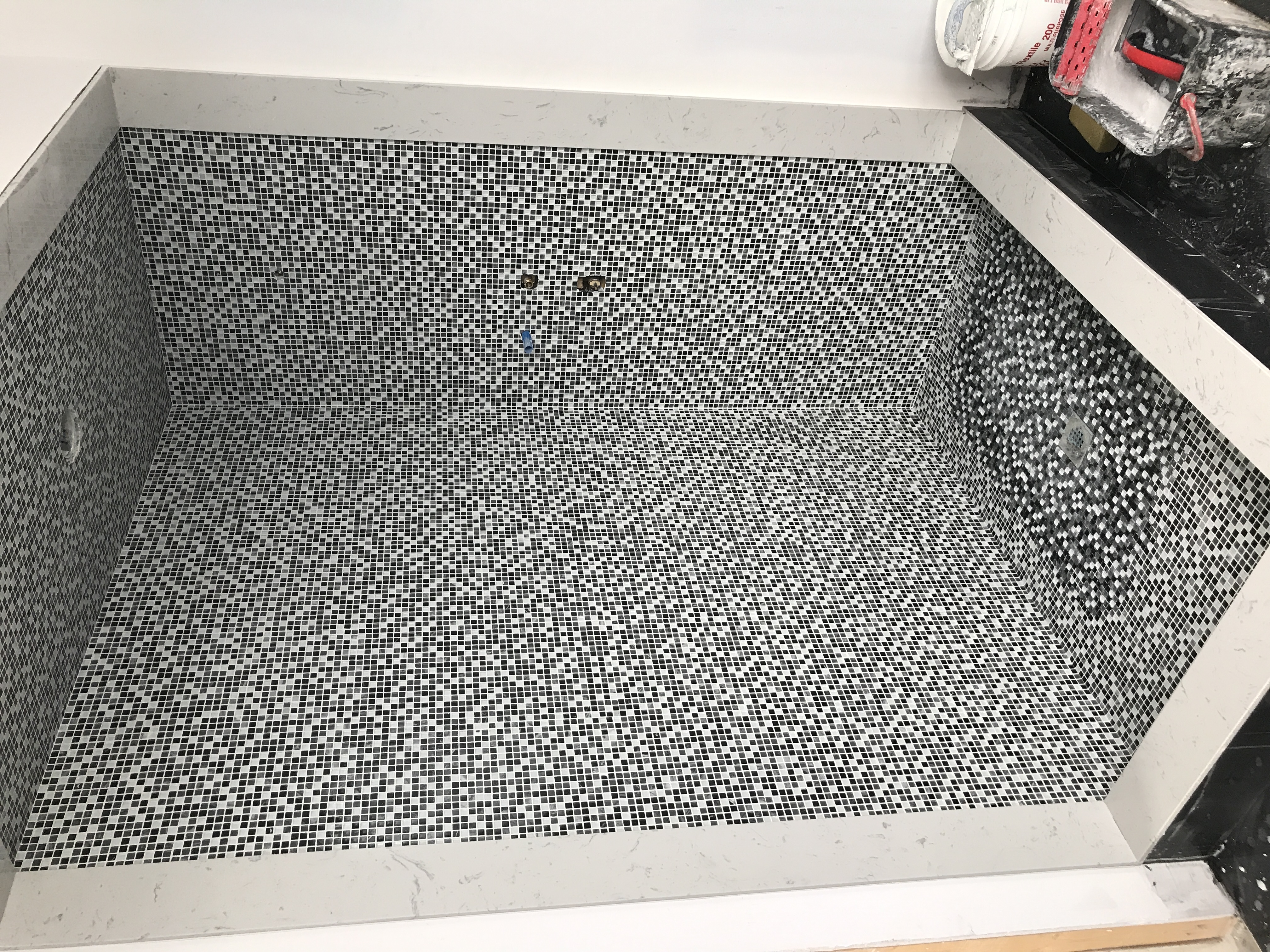 Wykończony brodzik z mozaiką czarno-białą, obramowany jasnym marmurem, widoczne odpływy liniowe i przyłącza wodne, w tle narzędzia glazurnicze.