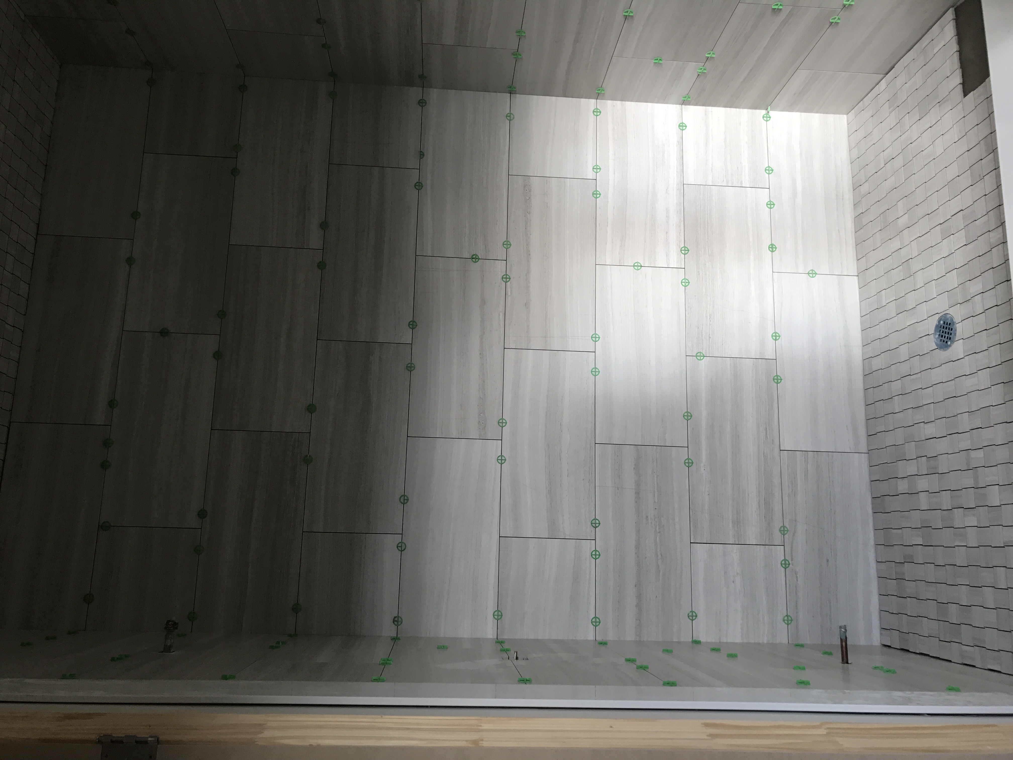 Wnętrze łazienki w trakcie układania płytek imitujących drewno, z użyciem krzyżyków dystansowych, widoczny odpływ liniowy i fragment ściany wykończonej mozaiką.