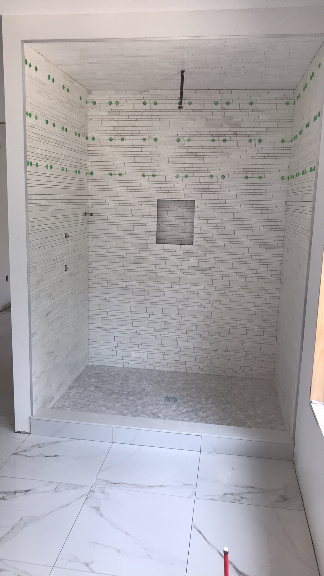 Wnętrze kabiny prysznicowej w trakcie wykańczania, z białymi, podłużnymi płytkami ułożonymi poziomo na ścianach, z widocznymi zielonymi krzyżykami dystansowymi, mała wnęka w ścianie, szara mozaika...