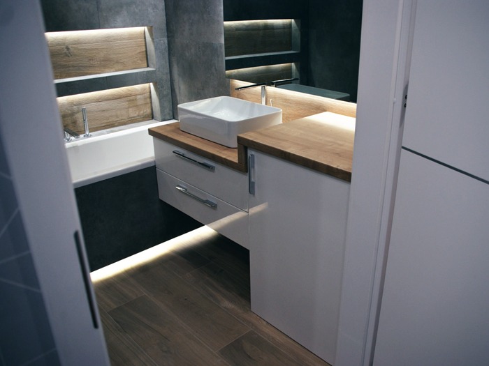 Nowoczesna łazienka w Bielsko-Białej z umywalką nablatową na białej szafce z drewnianym blatem, podświetlane wnęki w szarej ścianie oraz drewniana podłoga z listwą LED.