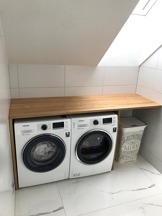 Dwie pralki Samsung pod drewnianym blatem w łazience na poddaszu, białe kafelki na ścianach i marmurowe na podłodze, obok kosz na pranie.