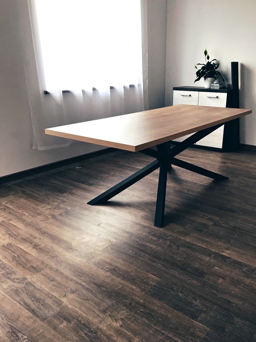 Jasny, drewniany stół z czarnymi, metalowymi nogami w kształcie gwiazdy, ustawiony na panelach podłogowych w pokoju z oknem i komodą w tle.