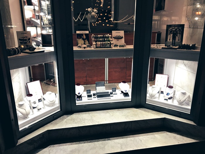 Witryna sklepowa z biżuterią, manekiny z naszyjnikami, bransoletki i kolczyki za szybą w kształcie trapezu, widoczne odbicia świateł i dekoracji świątecznych.