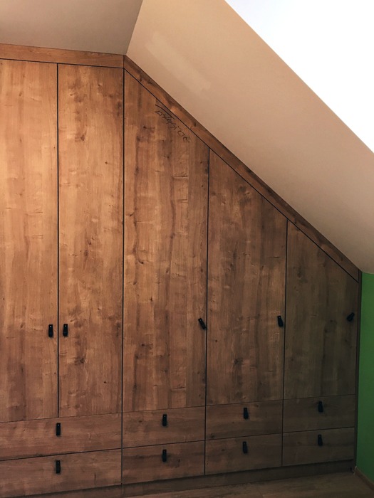 Drewniana szafa wnękowa z czarnymi uchwytami, dopasowana do skośnego sufitu, z widocznym fragmentem zielonej ściany.