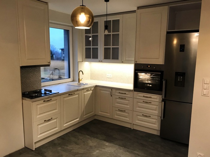 Jasna kuchnia z kremowymi szafkami z frezowanymi frontami, marmurowym blatem, złotą armaturą i designerską lampą wiszącą z przydymionego szkła.