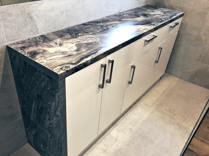 Szafka łazienkowa z białymi, lakierowanymi frontami i blatem imitującym marmur w odcieniach szarości i brązu, z podłużnymi, metalowymi uchwytami.