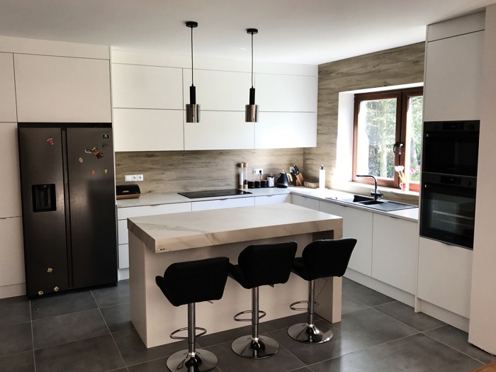 Nowoczesna kuchnia z białymi szafkami, wyspą z marmurowym blatem i trzema czarnymi hokerami, oświetlona wiszącymi lampami w metalowych obudowach.