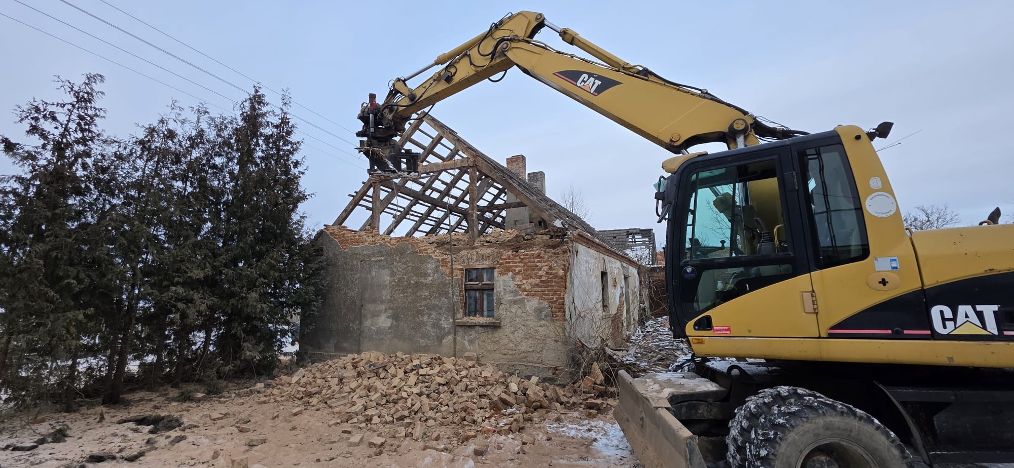 Żółta koparka CAT w trakcie rozbiórki ceglanego budynku z drewnianą konstrukcją dachu. Gruz na ziemi, drzewa w tle. Widoczne elementy zniszczonej elewacji.