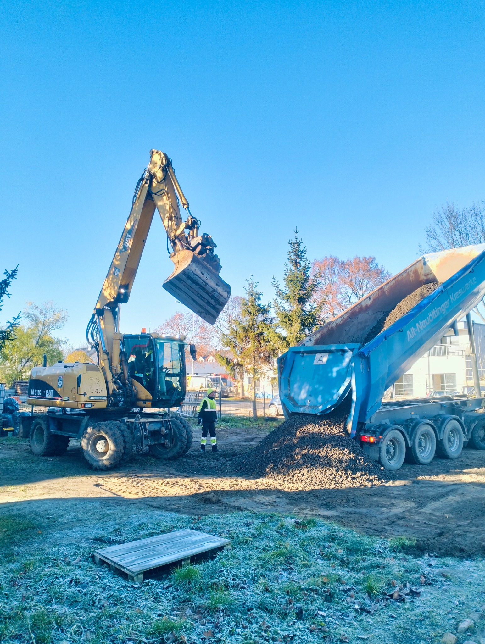 Żółta koparka CAT ładuje gruz do niebieskiej ciężarówki na tle błękitnego nieba. Na pierwszym planie oszroniona trawa i drewniana paleta. Praca w toku.