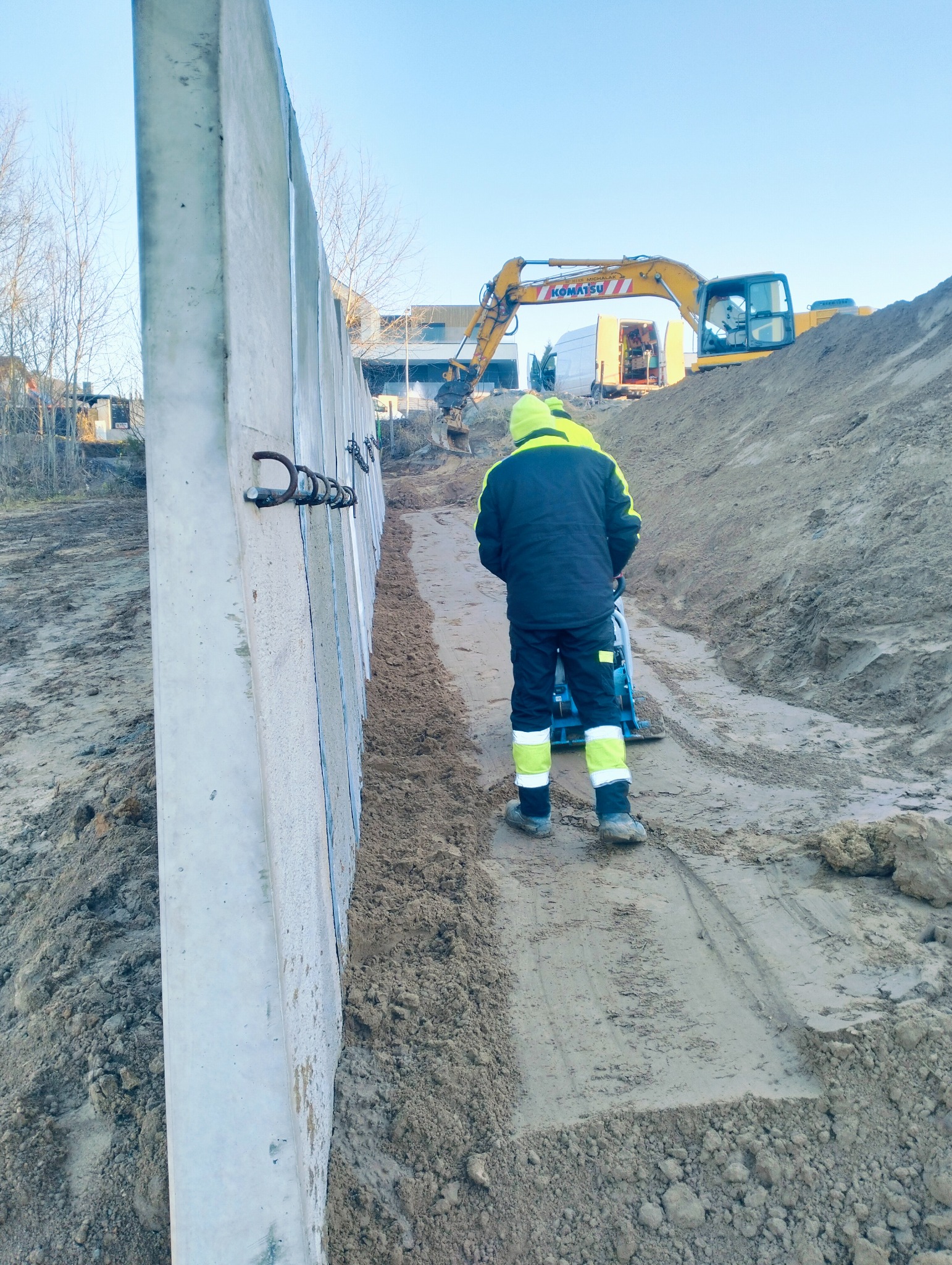 Prace ziemne: zagęszczanie gruntu wzdłuż betonowego muru oporowego. W tle koparka Komatsu i pracownicy w odblaskowych ubraniach. Widok z perspektywy pieszego.