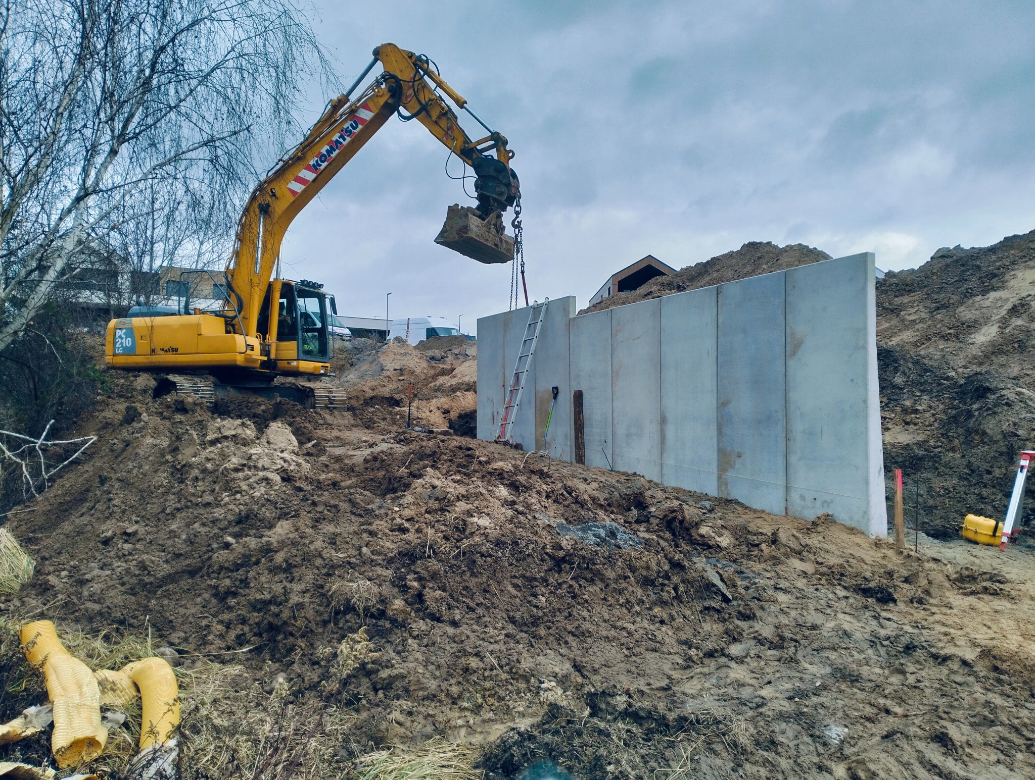 Żółta koparka Komatsu podnosi ziemię nad betonową ścianą oporową. W tle widoczne drzewa i budynki. Prace ziemne w toku, drabina oparta o ścianę.