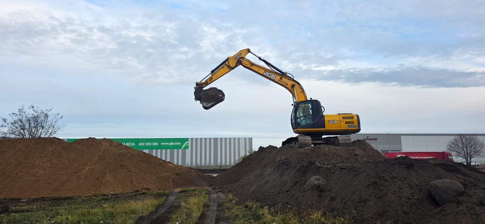 Żółta koparka JCB na hałdzie ziemi, z podniesionym ramieniem i łyżką. W tle budynek magazynowy z zielonym logo i pochmurne niebo.