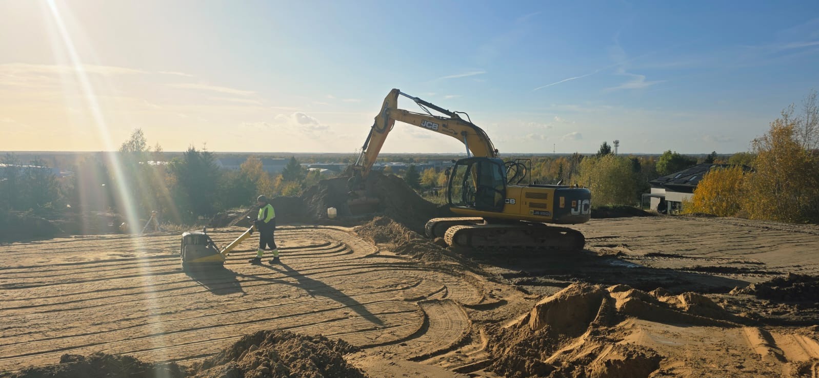 Żółta koparka JCB w trakcie niwelacji terenu pod budowę. W tle widoczny pracownik z zagęszczarką. Słoneczny dzień, błękitne niebo.