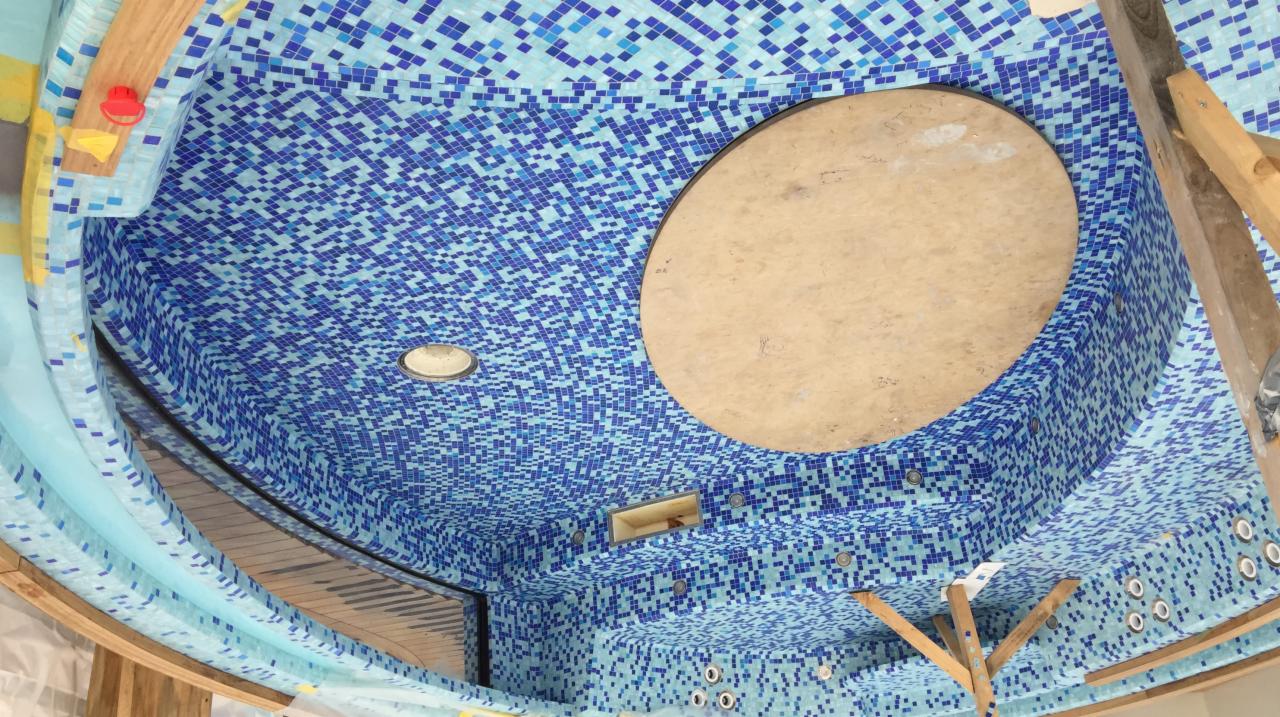 Mozaika z błękitnych kwadratów zdobi wnętrze wanny jacuzzi, z widocznym otworem na odpływ i drewnianą konstrukcją wspierającą, w trakcie prac wykończeniowych.
