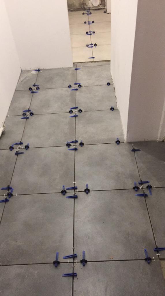 Szare płytki podłogowe w trakcie układania, z użyciem niebieskich klipsów poziomujących, widoczne fugi i fragment ściany