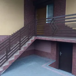 Brązowa, metalowa balustrada schodowa z geometrycznym wzorem, prowadząca do drzwi wejściowych budynku z ceglaną elewacją.