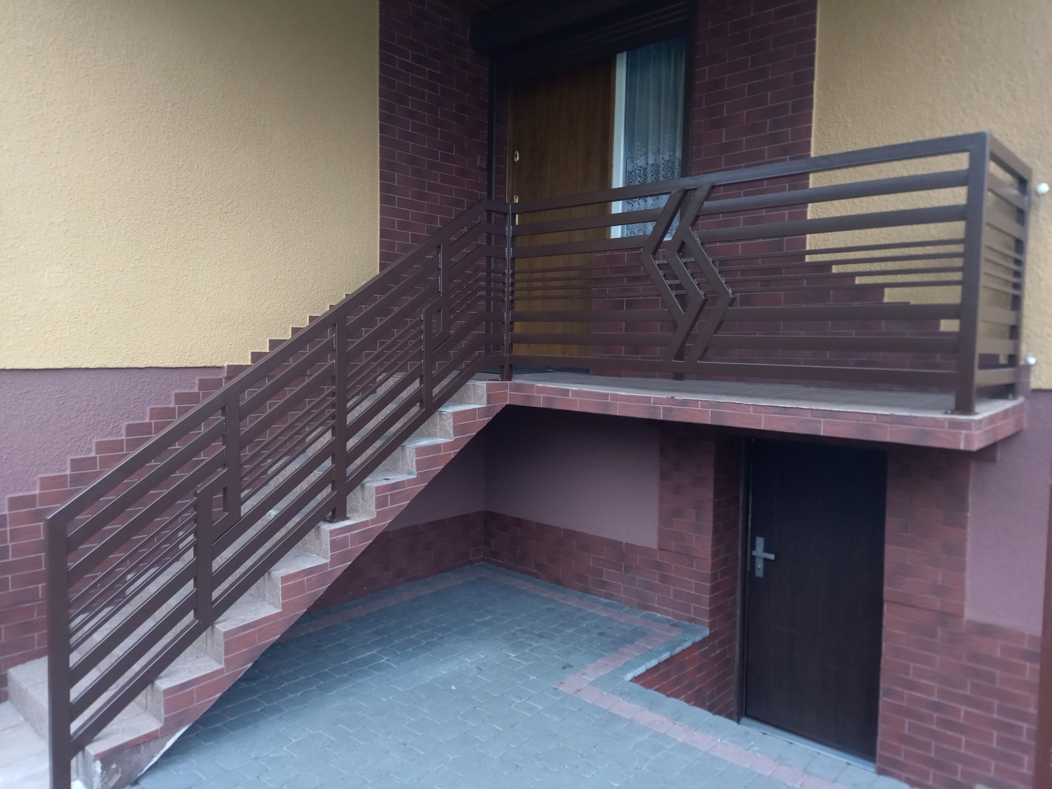 Brązowa, metalowa balustrada schodowa z geometrycznym wzorem, prowadząca do drzwi wejściowych budynku z ceglaną elewacją.