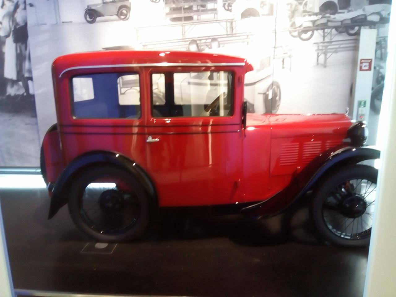 Czerwony, zabytkowy samochód z czarnymi błotnikami, stojący na czarnej powierzchni na tle ściany z czarno-białymi fotografiami starych samochodów.