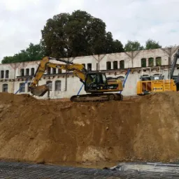 Żółta koparka i wiertnica Keller na placu budowy we Wrocławiu, na tle starego budynku z cegły i przygotowanego zbrojenia.