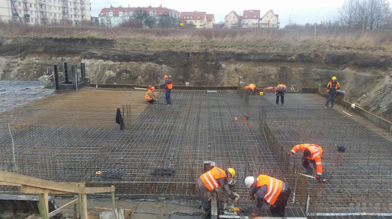Prace zbrojarskie na budowie: widok z góry na fundament z siatką zbrojeniową i pracujących robotników w kamizelkach odblaskowych, w tle budynki mieszkalne i wykop ziemny.