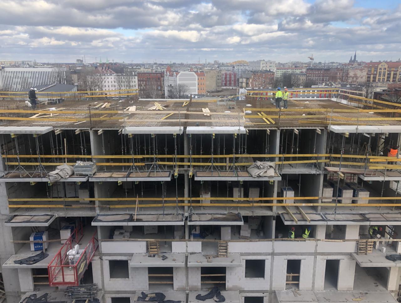 Wielopiętrowy budynek żelbetowy w trakcie budowy z widocznymi pracownikami w kamizelkach odblaskowych i kaskach, szalunkami, zbrojeniami i widokiem na panoramę miasta w tle.