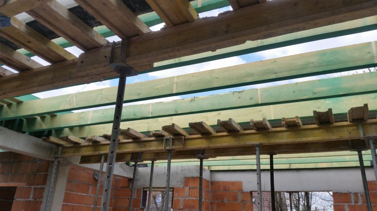 Wnętrze budynku w trakcie budowy z widocznym szalunkiem stropu podpartym metalowymi stemplami, ściany z czerwonej cegły dziurawki.