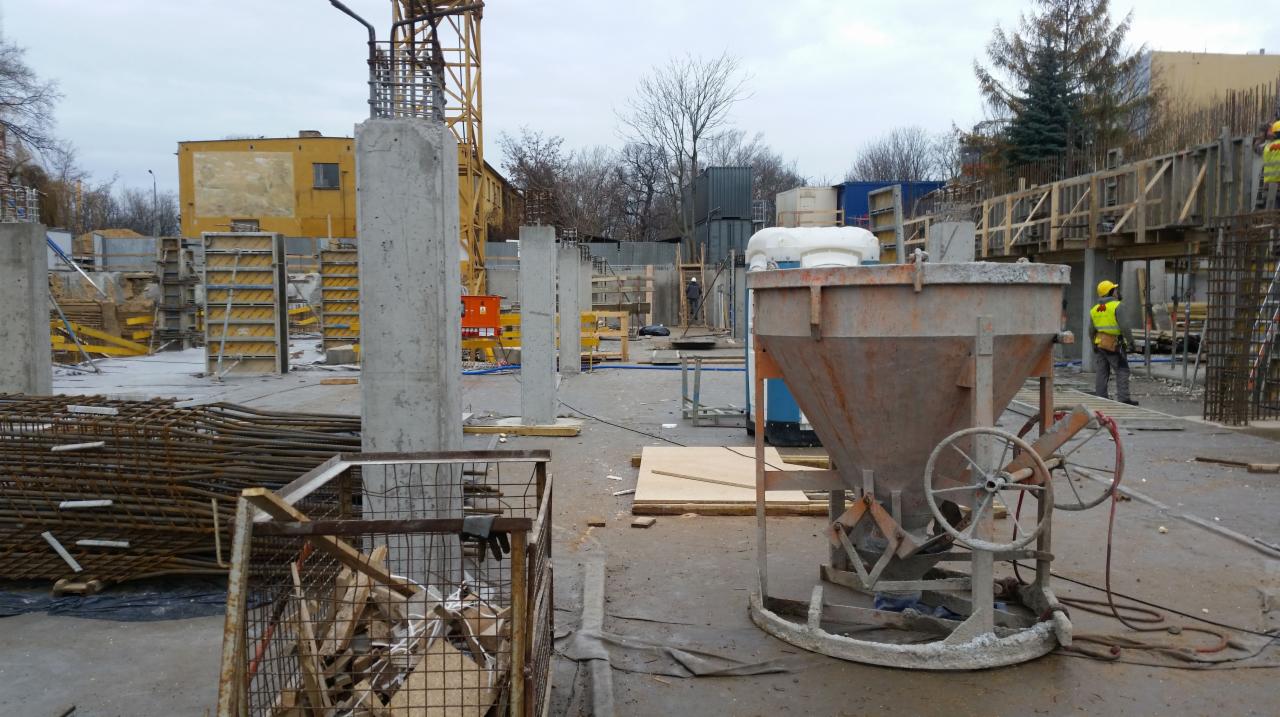 Plac budowy z widocznymi betonowymi słupami w szalunkach, zbrojeniami i lejkiem do betonu. W tle budynek w trakcie budowy i pracownik w żółtej kamizelce.