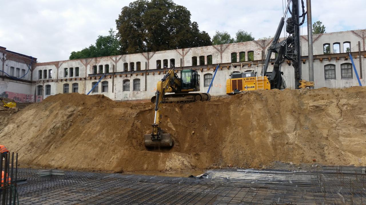 Żółta koparka i maszyna do palowania na placu budowy z widoczną siatką zbrojeniową i starym budynkiem w tle.