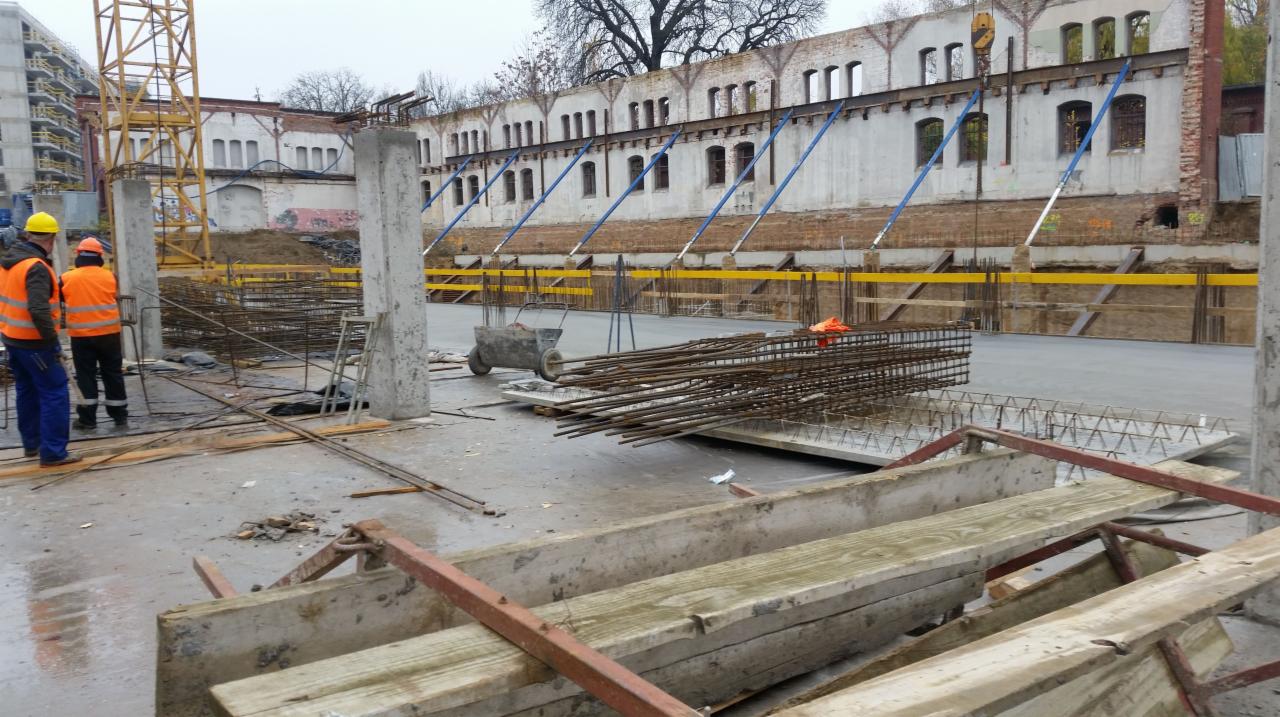 Plac budowy z widocznymi zbrojeniami, betonowymi słupami i fundamentami, w tle stara, odrestaurowywana kamienica z zabezpieczeniami.
