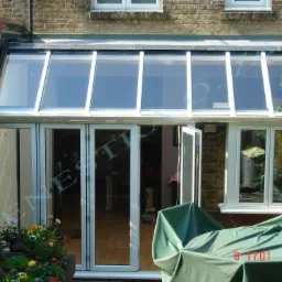 Zadaszony taras z białymi ramami okiennymi i składanymi drzwiami, widok z zewnątrz, donice z kwiatami na pierwszym planie, część mebli przykryta zieloną plandeką.