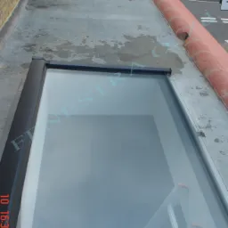 Widok z góry na płaski dach z dwoma oknami dachowymi w czarnych ramach, z widoczną drabiną wewnątrz pomieszczenia przez jedno z okien oraz ceglanym kominem i zaparkowanym samochodem w tle.