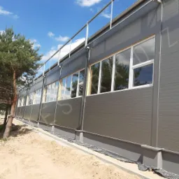 Szary budynek przemysłowy z rzędem białych okien PCV, widoczny fragment terenu z piaskiem i drzewo iglaste.