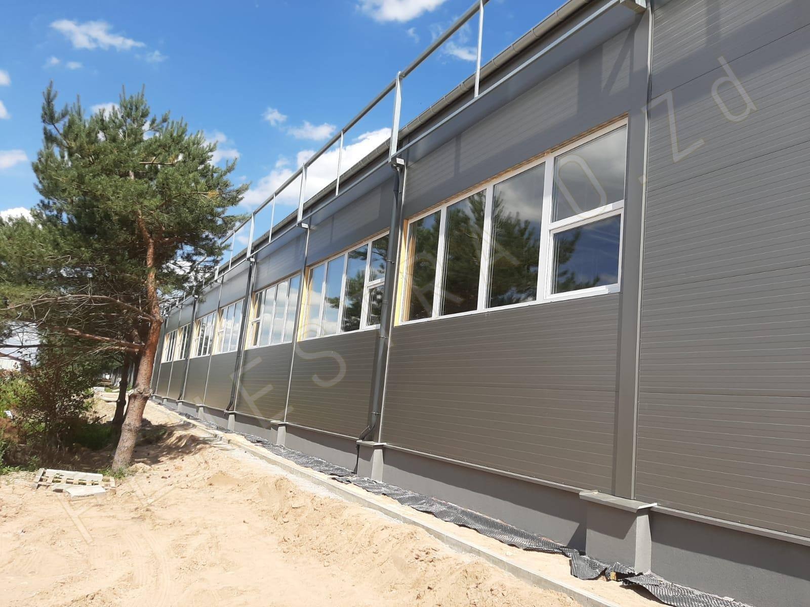 Szary budynek przemysłowy z rzędem białych okien PCV, widoczny fragment terenu z piaskiem i drzewo iglaste.
