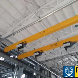 Dwie żółte suwnice pomostowe z wózkami jezdnymi zamontowane pod sufitem hali produkcyjnej.