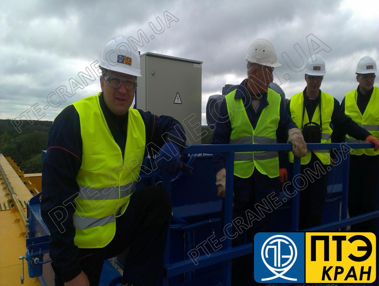 Grupa pracowników w kaskach i kamizelkach odblaskowych na platformie roboczej dźwigu, widoczne logo firmy PTE-CRANE