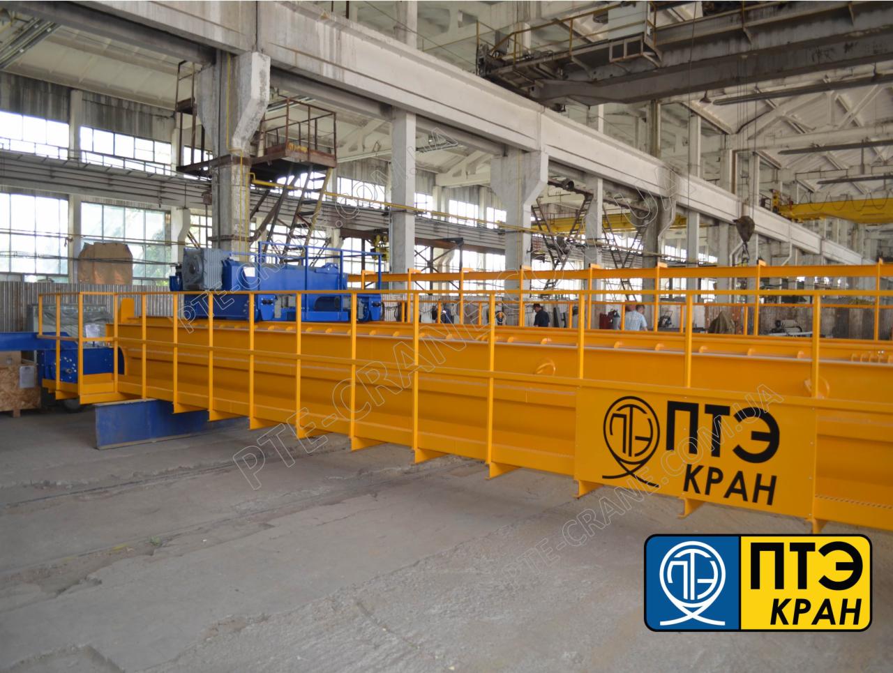 Żółto-niebieski suwnica pomostowa z widocznym logo producenta w hali przemysłowej z betonowymi kolumnami i oknami w tle.