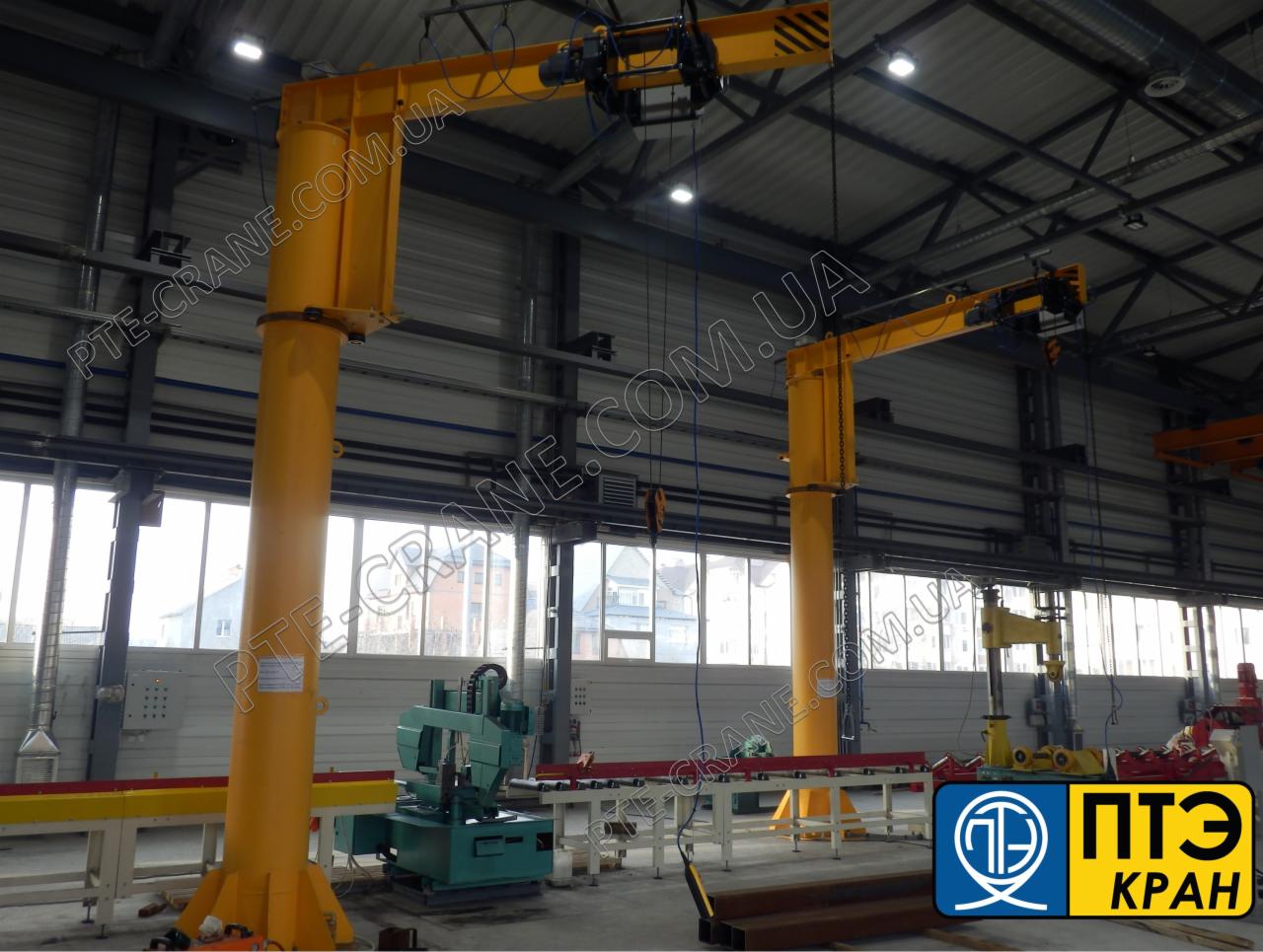 Żółty, obrotowy dźwig kolumnowy w hali produkcyjnej, z widoczną suwnicą pod stropem i urządzeniem do cięcia metalu w tle, z naniesionym znakiem wodnym na zdjęciu.