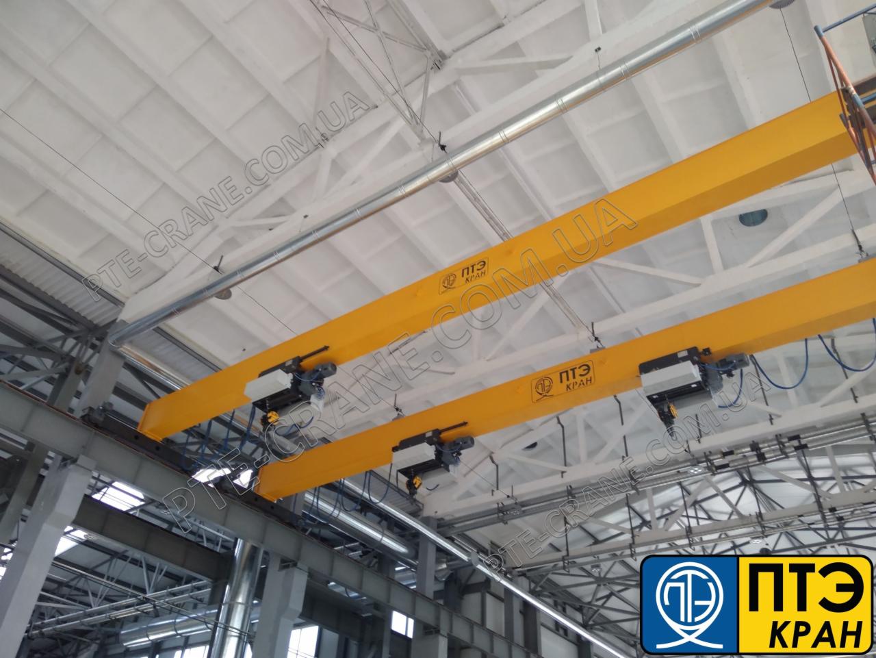Dwie żółte suwnice pomostowe z wózkami jezdnymi zamontowane pod sufitem hali produkcyjnej.