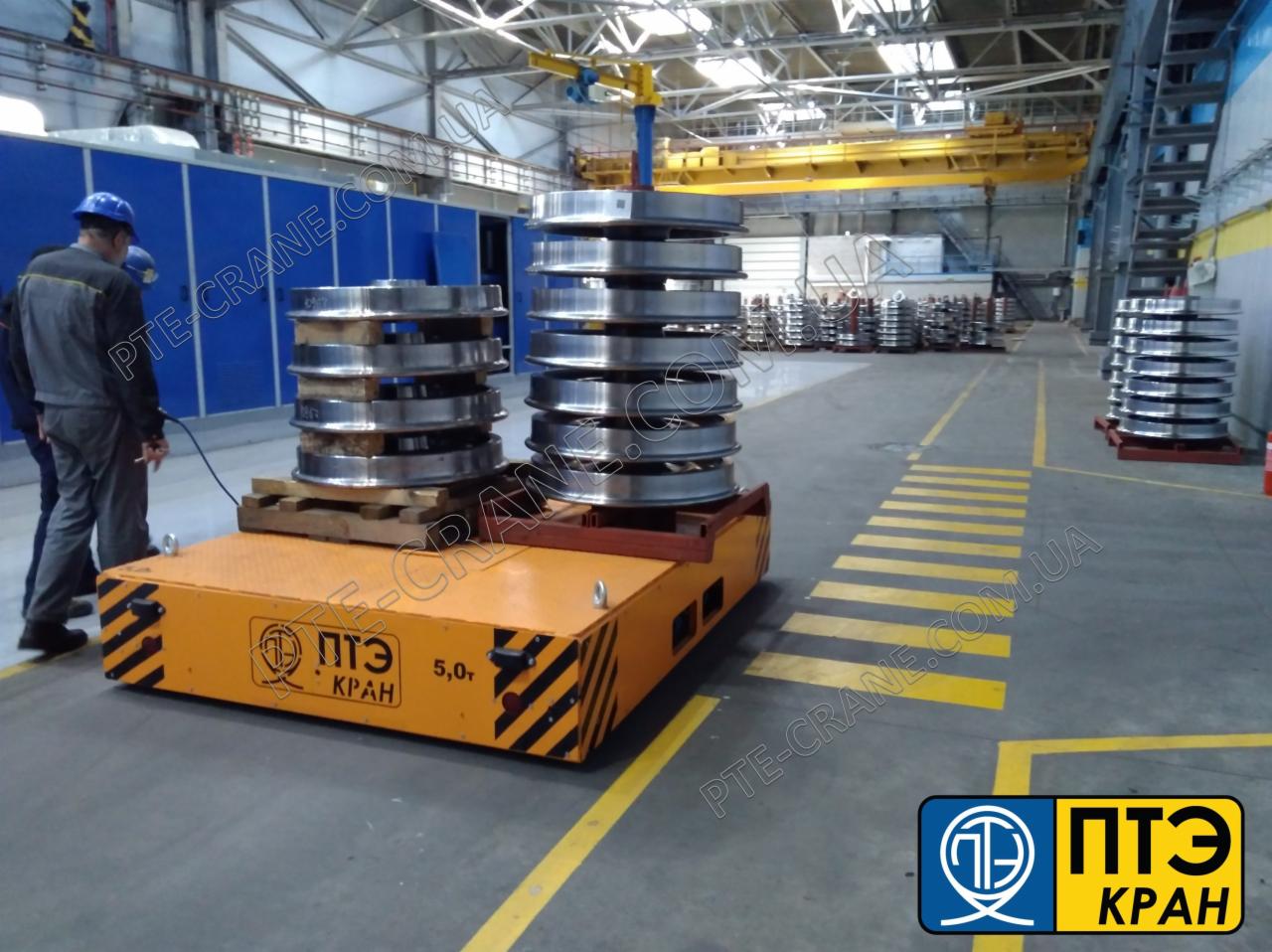 Wózek transportowy z ładunkiem metalowych obręczy w hali produkcyjnej w Warszawie, sterowany przez operatora w niebieskim kasku. Na drugim planie suwnica i regały z podobnymi elementami.