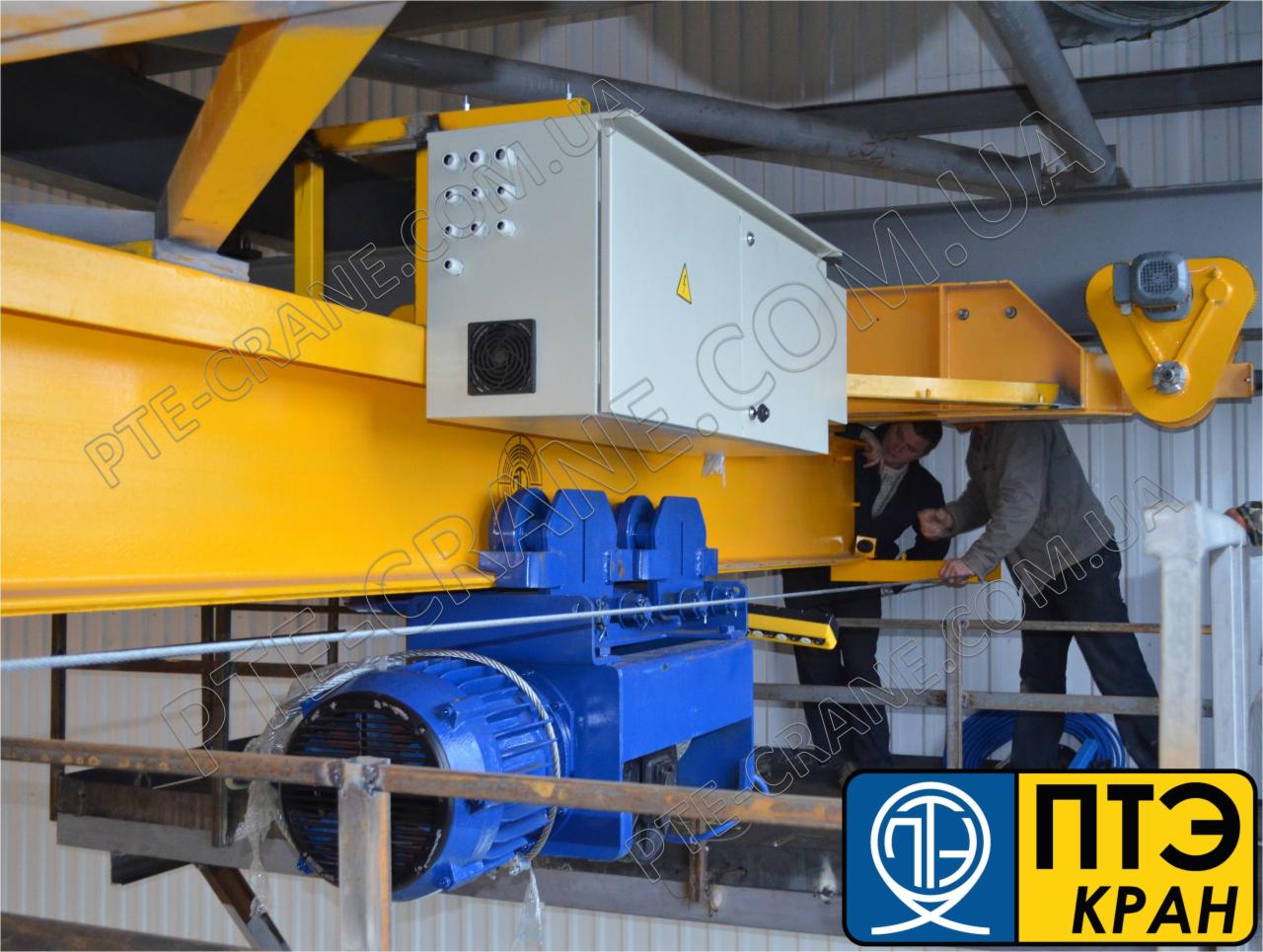 Żółto-niebieski suwnicowy dźwig podwieszany z elektrycznym silnikiem, widoczni dwaj pracownicy podczas instalacji lub konserwacji, w tle logo firmy.