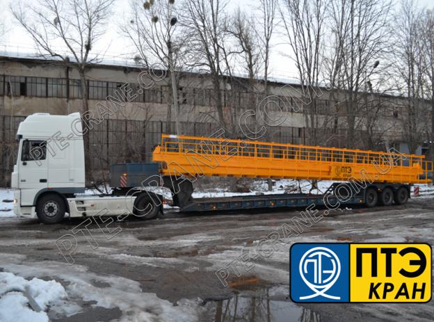 Ciężarówka z naczepą transportująca długi, żółty element konstrukcyjny przypominający pomost roboczy na tle szarego budynku przemysłowego.