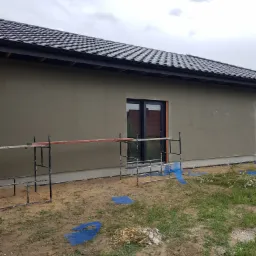 Świeżo otynkowana ściana domu jednorodzinnego z czarną dachówką, widoczne rusztowanie i folia ochronna na ziemi.