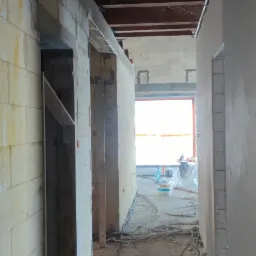 Wnętrze budynku w Bobrowicach w trakcie murowania ścian z bloczków, widoczne przewody na podłodze i surowy strop drewniany.