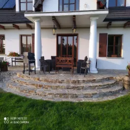 Kamienne schody tarasowe prowadzące do domu z białymi kolumnami i meblami ogrodowymi na górze, widok z trawnika.