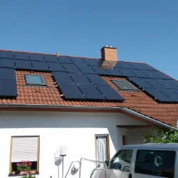 Dom jednorodzinny z czerwoną dachówką i zainstalowanymi panelami słonecznymi, widoczne okna dachowe i komin, jasne niebo w tle.