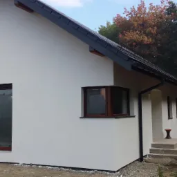 Jasny, otynkowany dom jednorodzinny z brązowymi oknami i ciemną dachówką, widok z boku na świeżo wyrównany teren przed budynkiem.