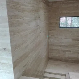 Wnętrze wykończone trawertynem: ściany i schody w odcieniach beżu i brązu, małe okno z widokiem na drzewa.